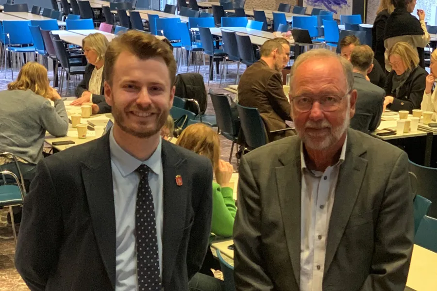 Et par menn i dress som sitter i et rom med andre mennesker
