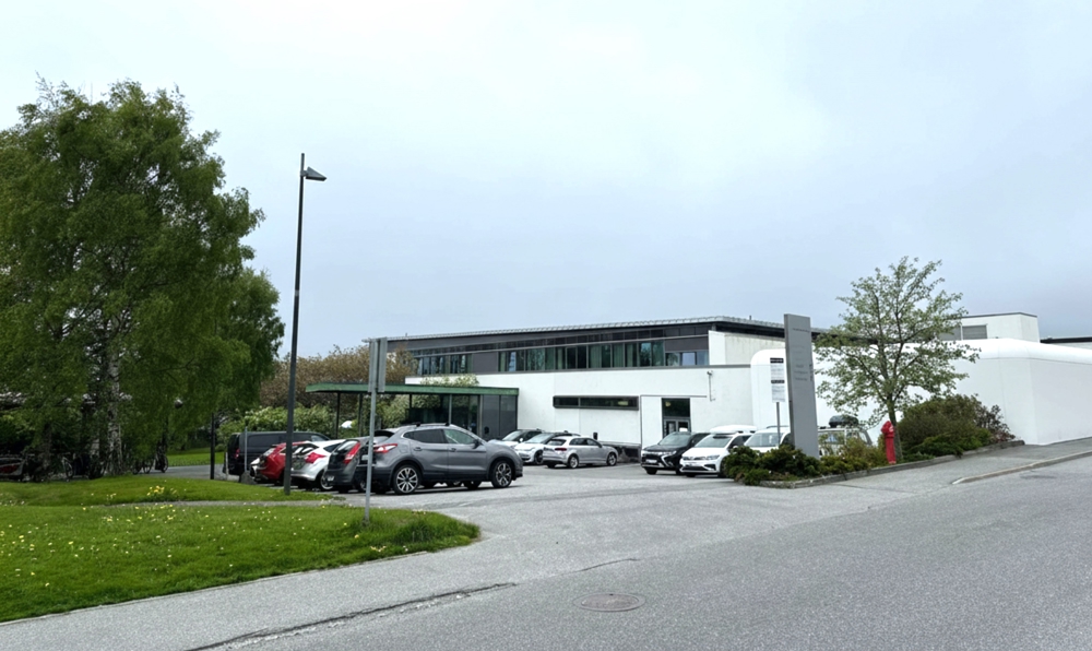 Eit kvitt bygg med overbygd inngangsparti, flatt tak og glassfasade i 2. etasje. Det står fleire bilar utanfor. 