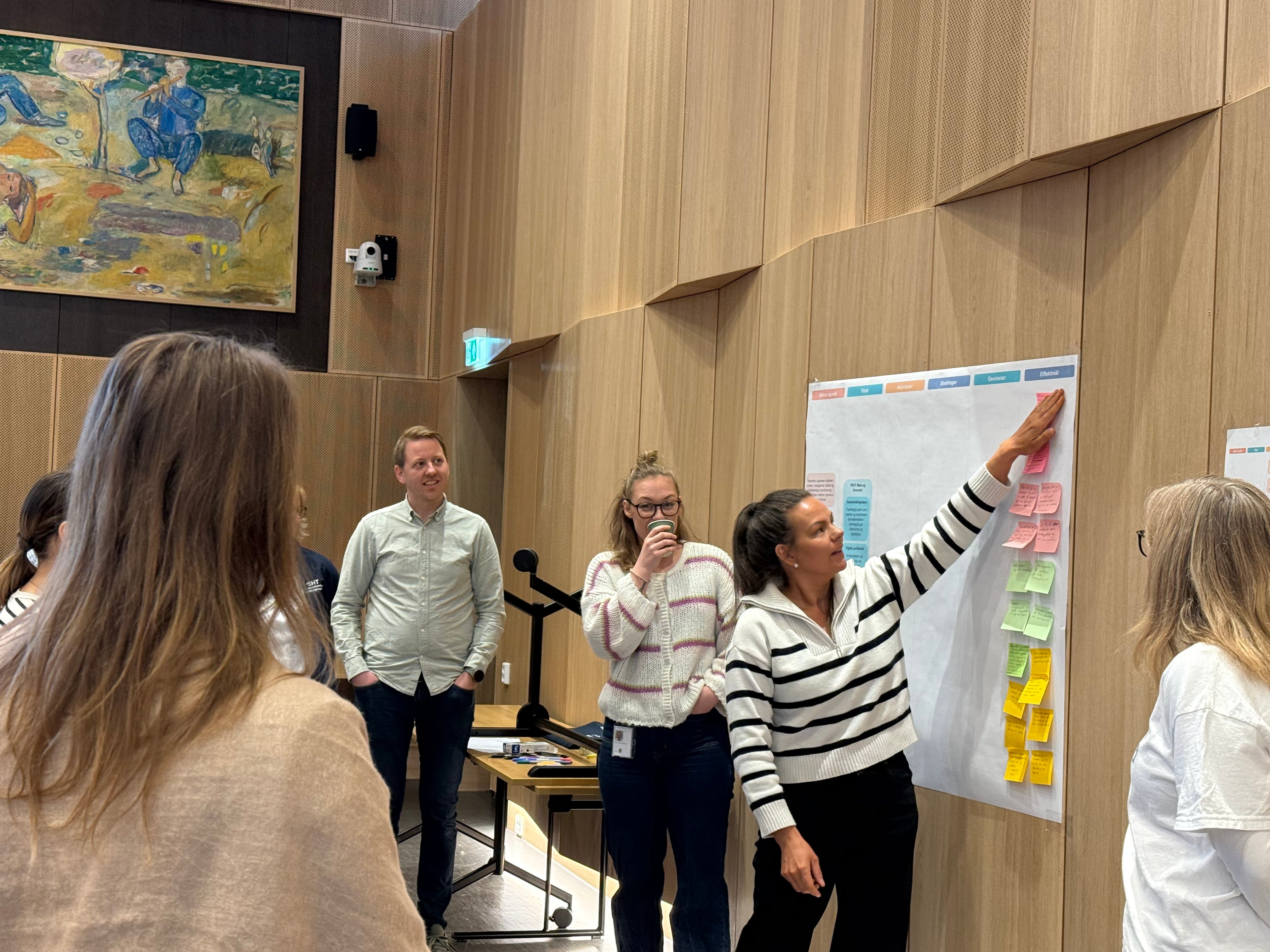 Ei dame som jobbar på ei tavle med post-it lappar, mens arbeidsgruppa står rundt. 