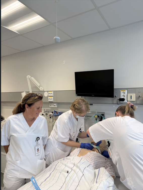 Tre personar i sjukehusuniform arbeider ved ei treningsdukke som ligg i ei sjukehusseng. Medisinsk utstyr er kopla til senga, og ein skjerm heng på veggen bak.