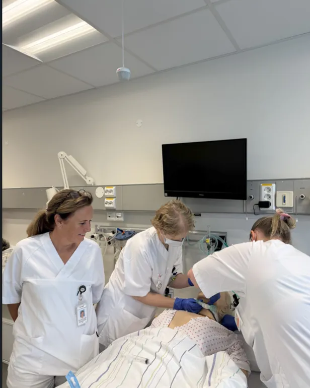 Tre personar i sjukehusuniform arbeider ved ei treningsdukke som ligg i ei sjukehusseng. Medisinsk utstyr er kopla til senga, og ein skjerm heng på veggen bak.
