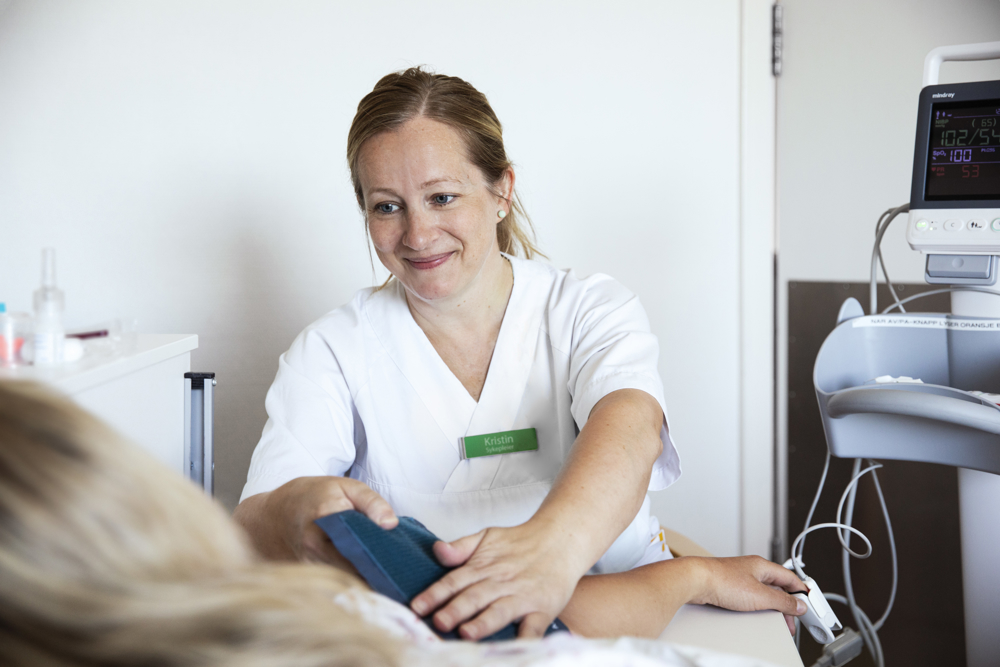 Ei smilande dame i kvitt arbeidstøy som måler blodtrykk på ein person som vi ser litt av