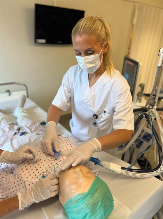 Ein person i sjukehusuniform og verneutstyr utfører ei prosedyre på ei treningsdukke som ligg i ei sjukehusseng. Respiratorutstyr og anna medisinsk utstyr står ved senga.
