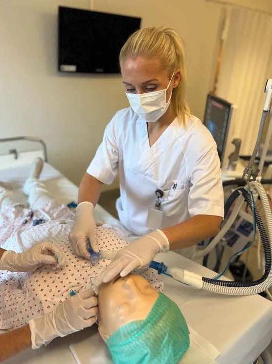 Ein person i sjukehusuniform og verneutstyr utfører ei prosedyre på ei treningsdukke som ligg i ei sjukehusseng. Respiratorutstyr og anna medisinsk utstyr står ved senga.