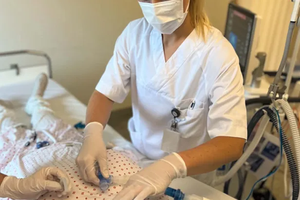 Ein person i sjukehusuniform og verneutstyr utfører ei prosedyre på ei treningsdukke som ligg i ei sjukehusseng. Respiratorutstyr og anna medisinsk utstyr står ved senga.