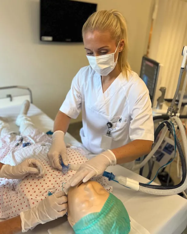 Ein person i sjukehusuniform og verneutstyr utfører ei prosedyre på ei treningsdukke som ligg i ei sjukehusseng. Respiratorutstyr og anna medisinsk utstyr står ved senga.