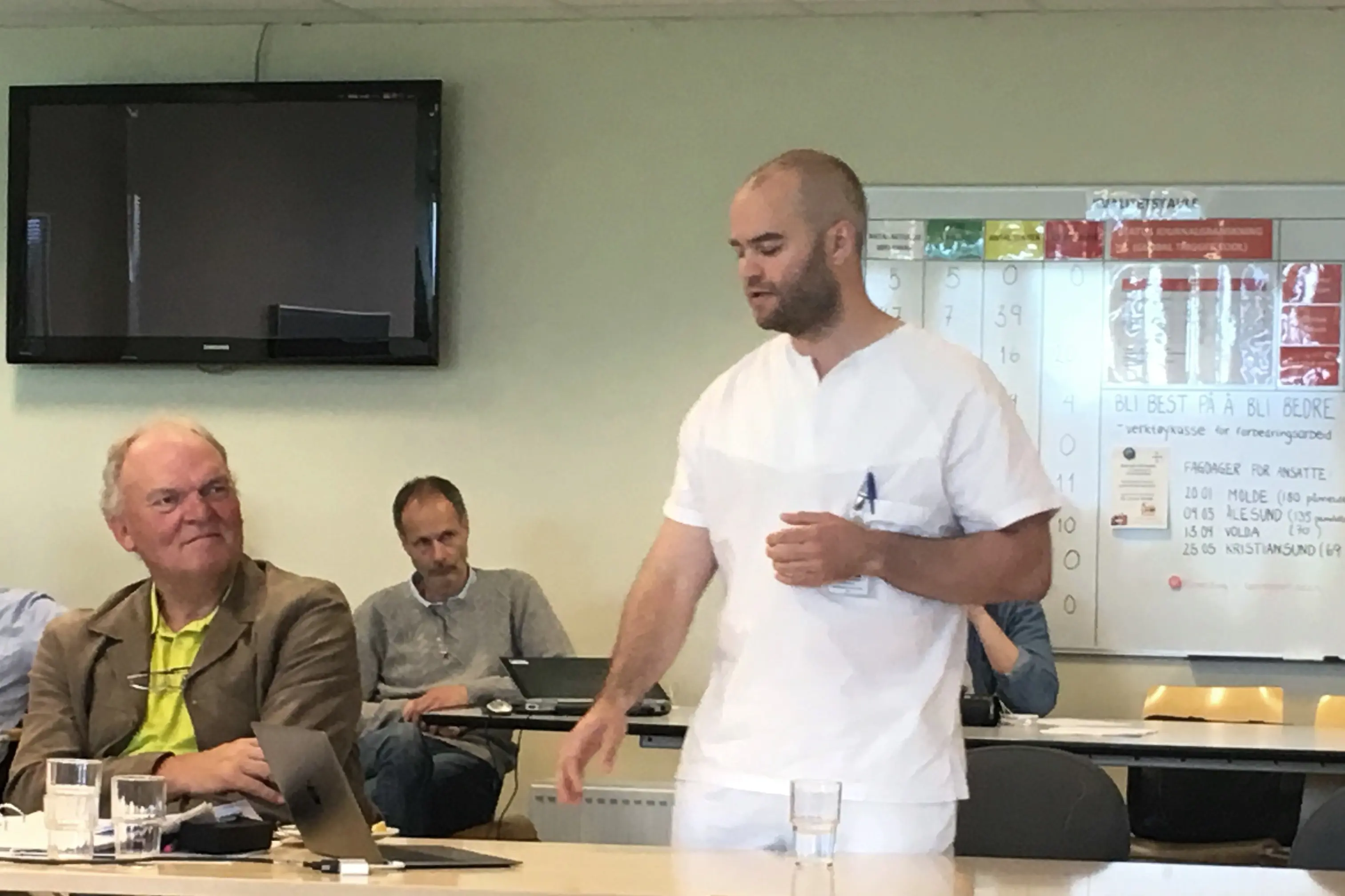 En mann som står foran et bord med andre menn som sitter ved det