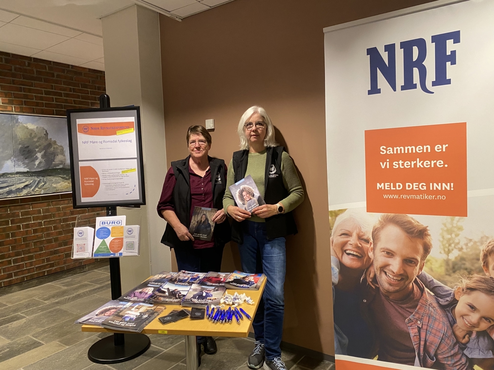 To kvinner som står i et rom bak et bord og holder hver sin brosjyre