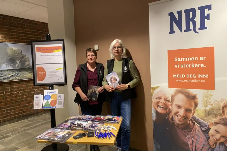 To kvinner som står i et rom bak et bord og holder hver sin brosjyre