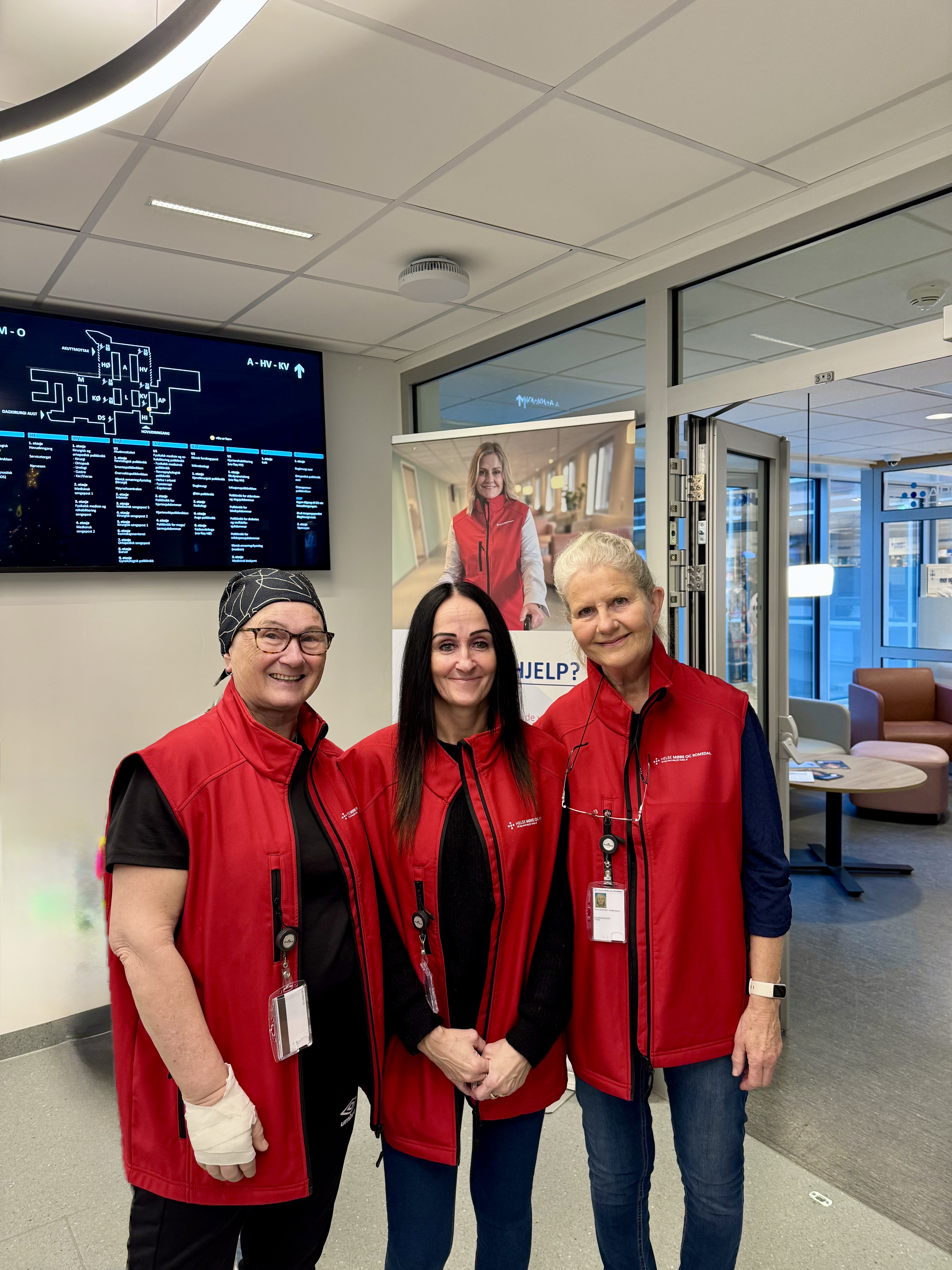 Tre smilande damer med raude vestar og id-kort. Dei står i hovudvestibylen på sjukehuset med ein informasjonsskjerm i bakgrunnen.