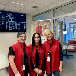 Tre smilande damer med raude vestar og id-kort. Dei står i hovudvestibylen på sjukehuset med ein informasjonsskjerm i bakgrunnen.