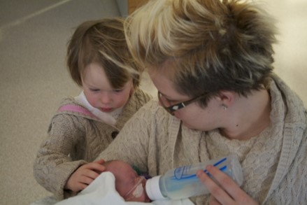 En kvinne som gir flaske til et nyfødt barn. En liten jente står ved siden av og holder en hånd på hodet til babyen.