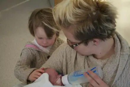 En kvinne som gir flaske til et nyfødt barn. En liten jente står ved siden av og holder en hånd på hodet til babyen.